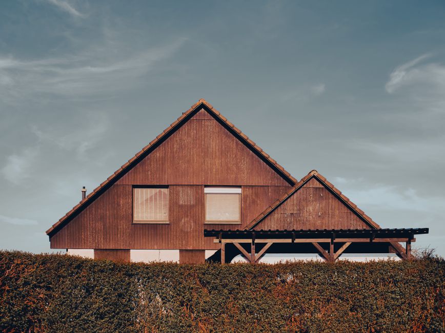 Aesthetic wooden house with triangular roof and shrubbery in front, under clear sky.