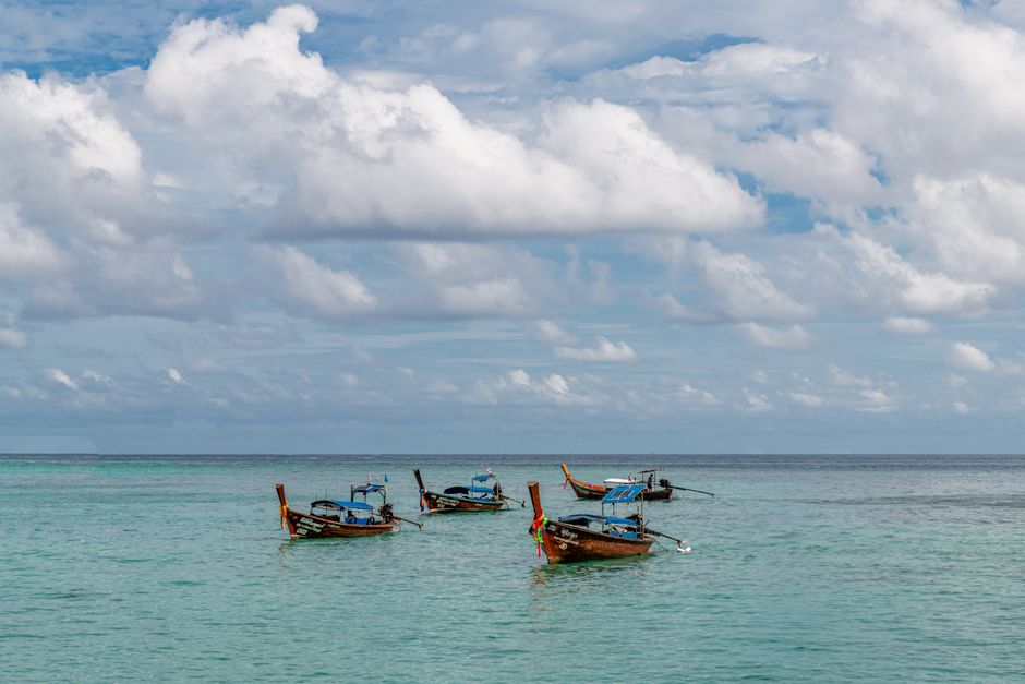 Traditional longtail boats floating on clear turquoise waters under a cloud-filled sky in Phuket, Thailand.