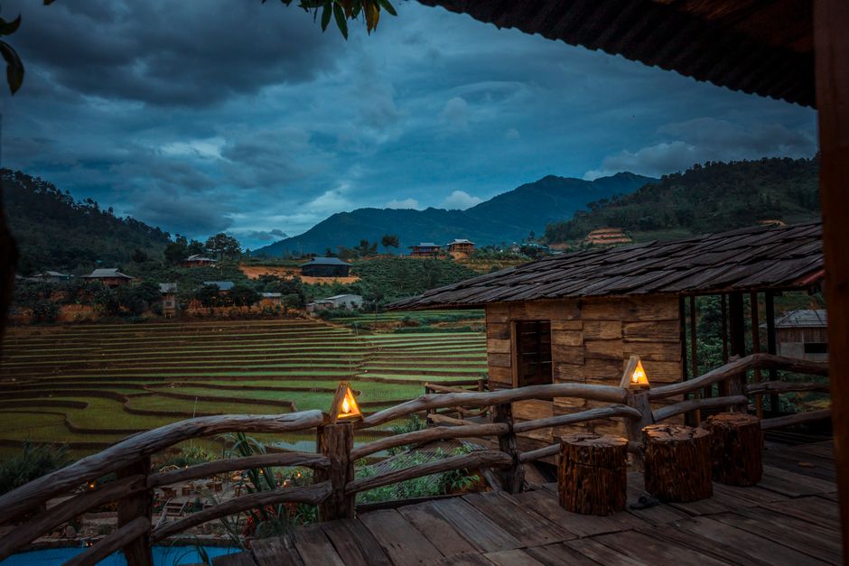 Scenic view of a rustic cabin with glowing lamps and rice terraces against a mountainous backdrop at dusk.