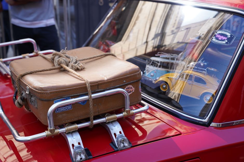 A vintage suitcase securely tied on a classic red car roof with retro stickers. Reflections in window.