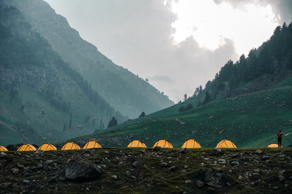 Scenic mountain campsite with illuminated tents and lone hiker under cloudy sky.