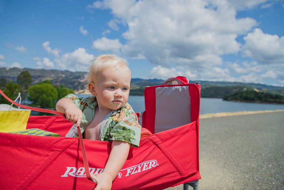 Adorable baby in a red wagon by a lake, enjoying a sunny outdoor day.