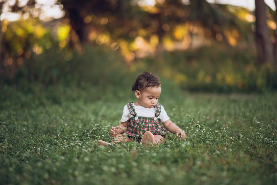 A toddler sits in the grass, surrounded by nature, exploring the outdoors.