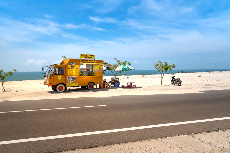 A vibrant yellow food truck by a sandy beach, with a clear blue sky.