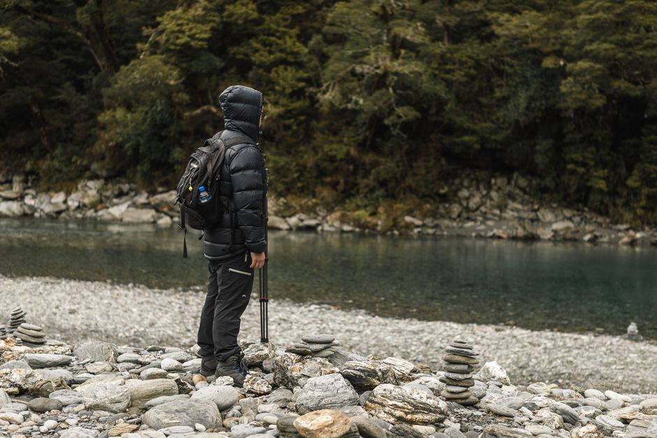 Hiker by a peaceful river, bundled for winter adventure in lush nature.