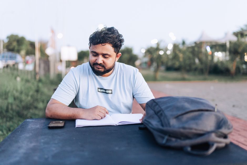 A man in a t-shirt studies an open notebook outdoors at a park table during the day.