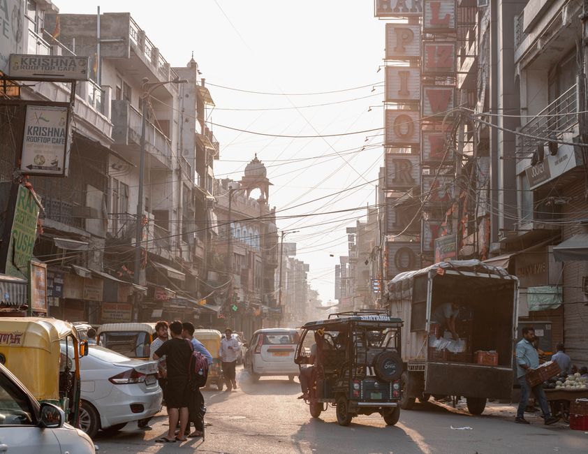 A busy street in Delhi with people, vehicles, and shops under a hazy sky.