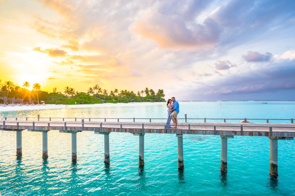 A couple embraces on a wooden pier with palm trees and a colorful sunset backdrop.