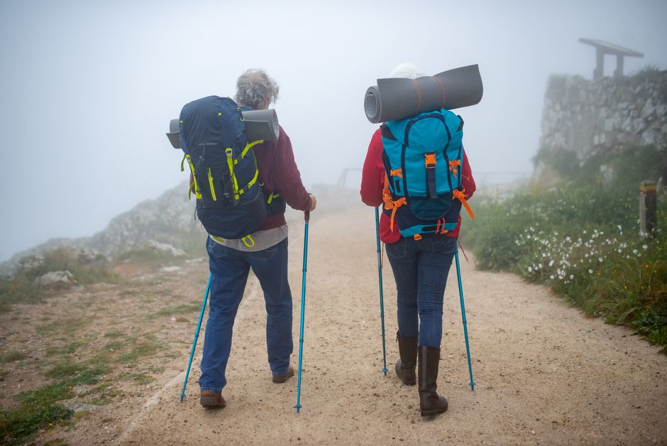 Elderly couple hiking on a foggy trail in Portugal, equipped with backpacks and walking poles.