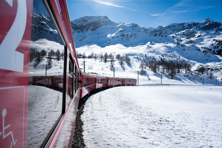 A picturesque red train curves through snowy alpine terrain under a clear blue sky.