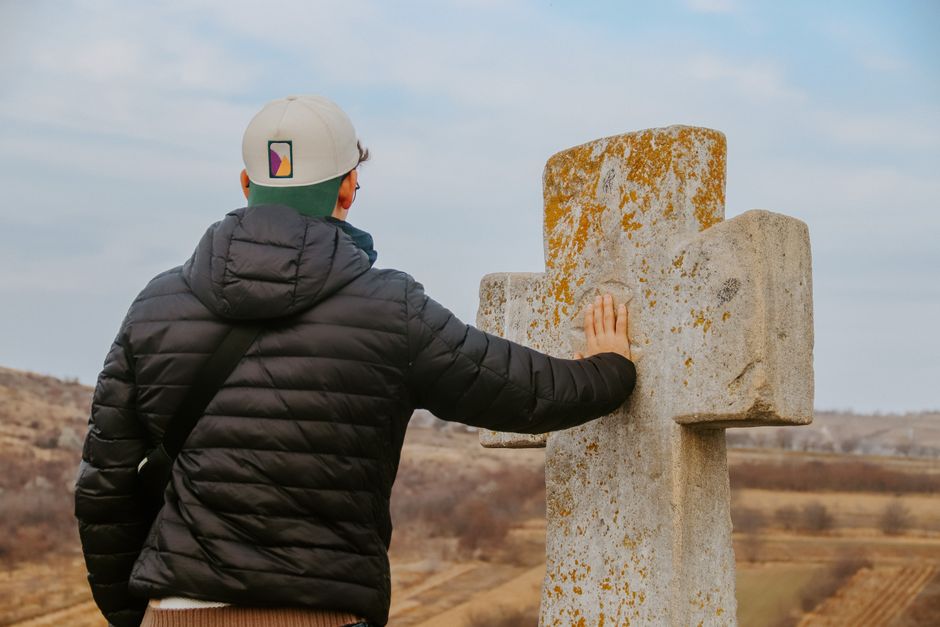 A man in a black jacket and cap touches a weathered stone cross in an open rural landscape.