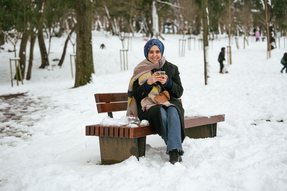 Smiling woman with smartphone seated on a snowy park bench in winter.