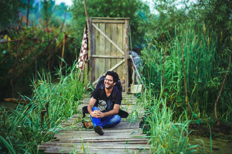 Man sitting and laughing on a boardwalk surrounded by wild grass, exuding a sense of adventure and relaxation.
