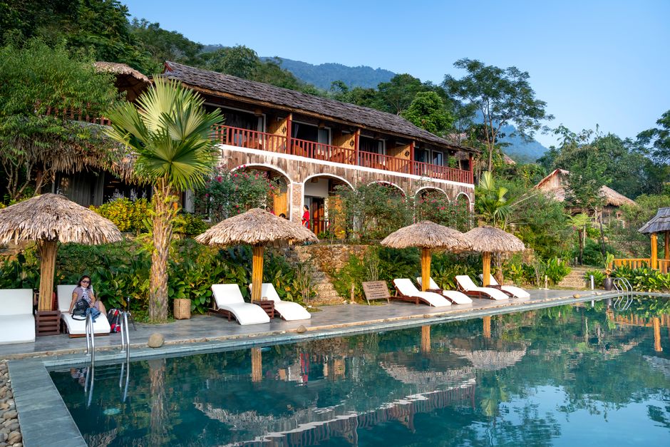 Swimming pool with blue water surrounded with sunbeds and straw umbrellas near small tropi