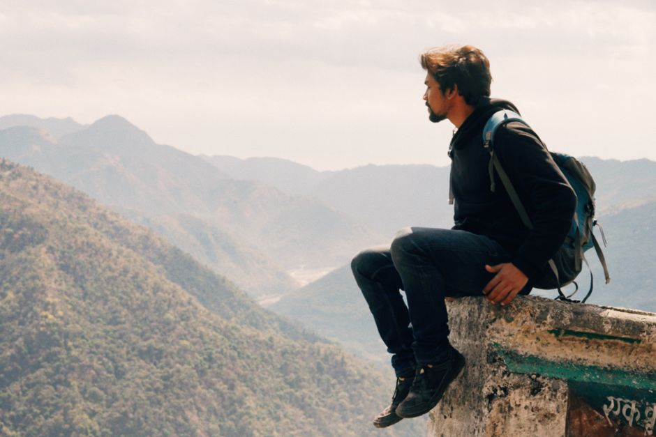 Side view of traveling man with backpack sitting on high edge with landscape of mountains in haze