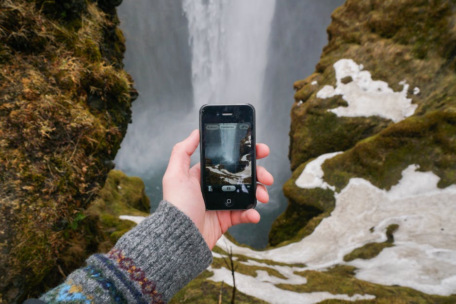Hand holding a smartphone photographing a waterfall amidst mossy rocks.