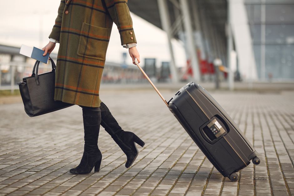 Crop young female traveler in trendy plaid coat and high boots with luggage and documents strolling on pavement near contemporary building of airport during cold season
