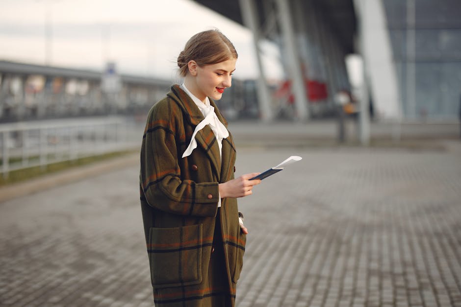 Smiling female passenger wearing trendy plaid coat and white blouse checking passport and ticket standing on pavement near contemporary building of airport
