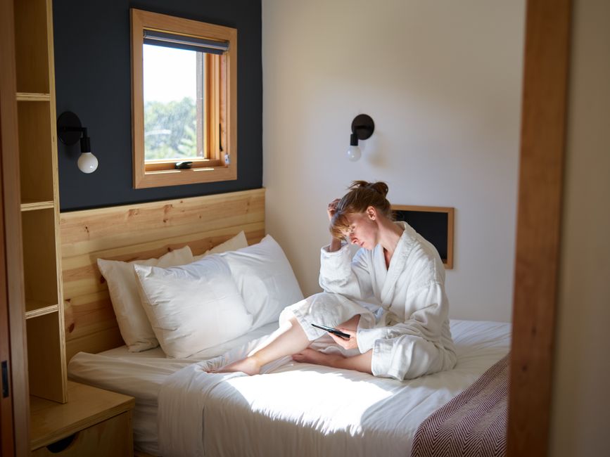 Full length barefoot focused female in white warm bathrobe chilling on cozy bed with ebook in modern bedroom on sunny day
